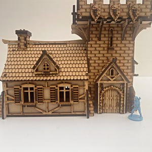 Op de afbeelding: Een houten model van een middeleeuws gebouw met een toren en een aangebouwd huis. De toren heeft een bakstenen gevel en een puntdak. Het huis heeft een pannendak en een deur met een decoratieve boog. Het model is geschilderd in tinten bruin en beige.