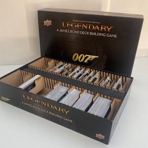 May include: A black box for the Legendary: A James Bond Deck Building Game, with the lid open. The box contains card decks and dividers. The text "LEGENDARY" and "007" are in gold. The box is made of wood.