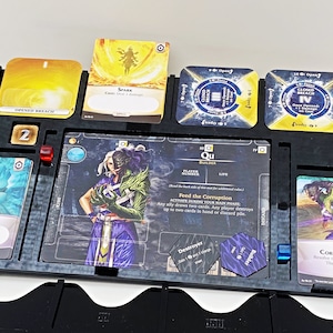 May include: A black game board with a green and purple character card, a blue and white card, a yellow card, and a red die. The character card has the text "Qu" and "Feed the Corruption".