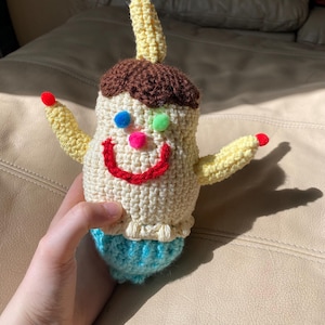 Puede incluir: Muñeca de ganchillo hecha a mano con un diseño caprichoso. La muñeca presenta un cuerpo color crema, cabello castaño y una falda azul. Tiene brazos amarillos y una protuberancia amarilla alta con puntas rojas. La cara tiene una sonrisa roja y ojos de pompón coloridos.