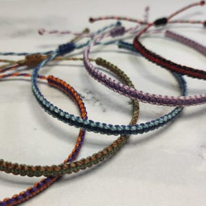 May include: Five colourful friendship bracelets made of woven string. The bracelets are in shades of blue, purple, red, green, and orange.