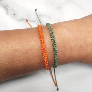 May include: Two friendship bracelets, one orange and one green, are tied around a wrist.