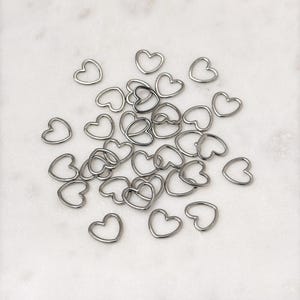 May include: A large group of silver heart-shaped jump rings. The rings are scattered on a white surface.