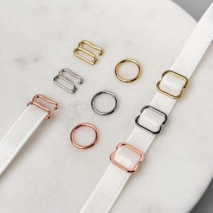 Puede incluir: Surtido de reguladores de tirantes de sujetador de metal en acabados dorado, plateado y oro rosa. Los reguladores son de diferentes formas, incluyendo cuadradas y rectangulares.