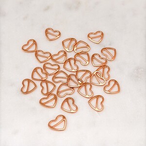 May include: A group of rose gold heart-shaped bra strap adjusters. The adjusters are scattered on a white marble surface.