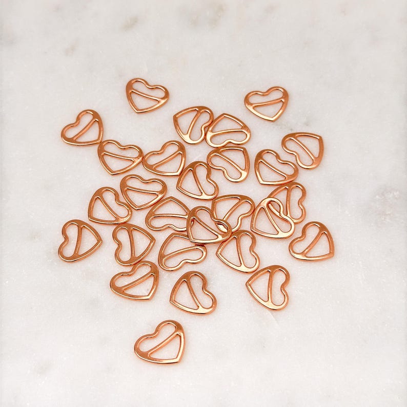 May include: A group of rose gold heart-shaped bra strap adjusters. The adjusters are scattered on a white marble surface.