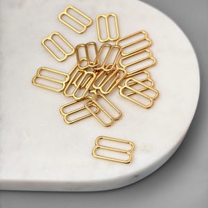 May include: A pile of gold-toned metal slider buckles. The buckles are oval-shaped with two holes on each side.