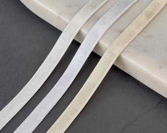 Könnte beinhalten: Nahaufnahme von drei elastischen Bändern in Weiß-, Silber- und Beigetönen. Die Bänder sind schräg über den Rahmen angeordnet und haben eine strukturierte Oberfläche. Der Hintergrund ist eine marmorähnliche Oberfläche und eine dunkelgraue Oberfläche.