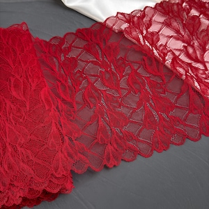 May include: Close-up of a vibrant red lace fabric with a delicate leaf pattern. The lace is folded over, revealing its intricate design and texture. The fabric is likely used for apparel or craft projects.