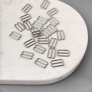 Peut inclure: Gros plan sur un tas de boucles coulissantes en m&eacute;tal argent&eacute;. Les boucles sont dispos&eacute;es sur une surface en marbre blanc.