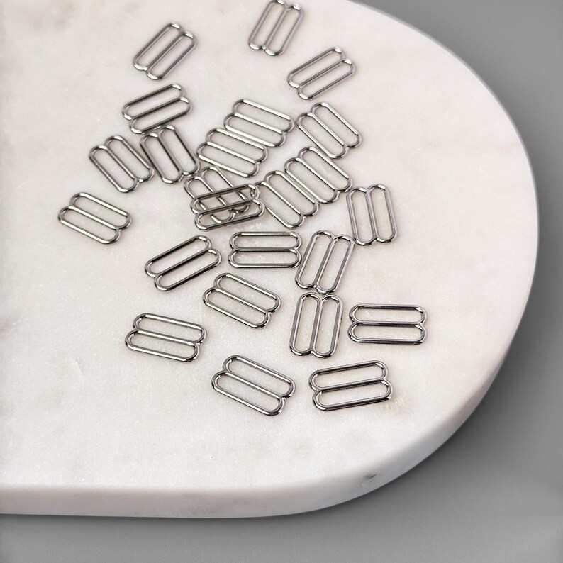 Peut inclure: Gros plan sur un tas de boucles coulissantes en m&eacute;tal argent&eacute;. Les boucles sont dispos&eacute;es sur une surface en marbre blanc.
