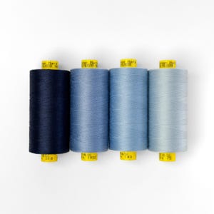 Puede incluir: Cuatro carretes de hilo en tonos de azul, desde azul oscuro a azul claro. Los carretes están etiquetados con el nombre de la marca "Gütermann Mara" y el tipo de hilo "Polyester 100".