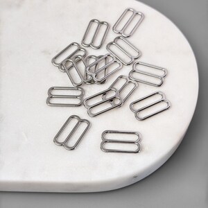 May include: A pile of silver metal slider buckles, commonly used for adjusting the length of straps on clothing or bags.