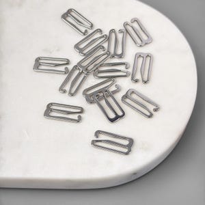May include: A close-up of a group of silver bra strap extenders scattered on a white marble surface.