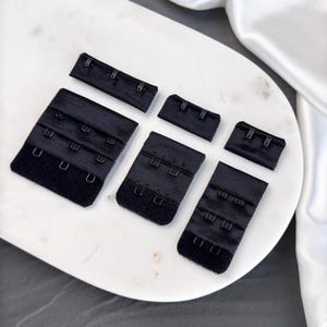 May include: A selection of black bra hook and eye closures in different sizes. The closures are made of fabric and metal, with various hook and eye configurations. They are displayed on a white, marble-effect surface.