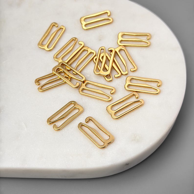 May include: A close-up of a pile of gold-colored metal bra strap extenders. The extenders are in the shape of a rectangle with two loops on each side.