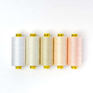 Könnte beinhalten: Fünf Garnrollen in verschiedenen Weiß- und Pfirsichtönen. Die Rollen sind mit "Gütermann Mara 100% Polyester" beschriftet und haben Nummern.