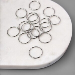 May include: A collection of silver jump rings, scattered on a white marble surface. The rings are various sizes and are perfect for jewelry making.