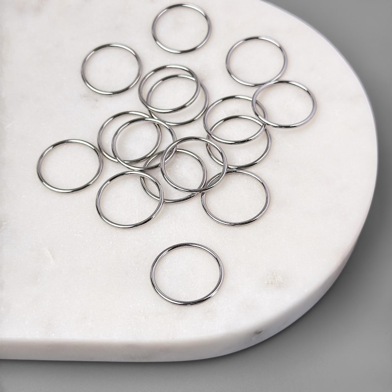 May include: A collection of silver jump rings, scattered on a white marble surface. The rings are various sizes and are perfect for jewelry making.