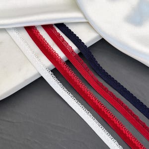 Può includere: Primo piano di quattro nastri elastici bianchi, rossi e blu navy. Ogni nastro ha un bordo smerlato. I nastri sono disposti diagonalmente su uno sfondo bianco e grigio.