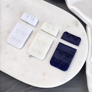 Könnte beinhalten: Vier BH-Verlängerungen, zwei weiße und zwei marineblau. Die Verlängerungen haben drei Haken und Ösen.