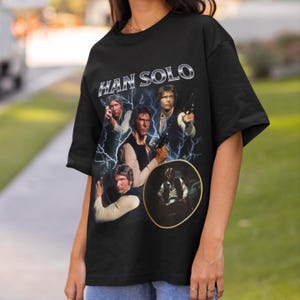 May include: A black short-sleeve t-shirt featuring a graphic print of Han Solo from Star Wars. The design includes multiple images of the character, blue lightning effects, and the text HAN SOLO in a metallic silver font across the top.