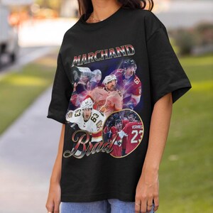 以下が含まれることがあります： 黒いTシャツ。ホッケーをテーマにしたデザインで、メタリックフォントで「Marchand」という名前、ホッケー選手のコラージュ、筆記体フォントで「Brad」という名前が特徴です。