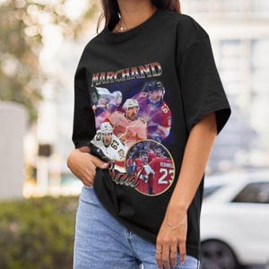 以下が含まれることがあります： 黒いTシャツ。ホッケーをテーマにしたデザインで、ホッケー選手のコラージュ、銀色の「MARCHAND」の文字、赤と白のジャージを着た選手の画像が特徴です。63と23の数字が見えます。ヴィンテージスタイルのデザイン。