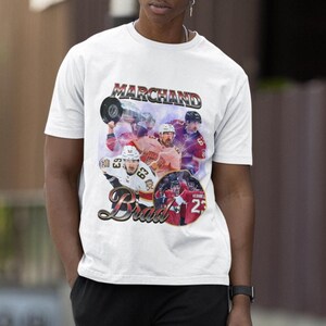 以下が含まれることがあります： 白のTシャツ。ヴィンテージスタイルのグラフィックで、ホッケー選手、上部に「Marchand」、下部に「Brad」の文字。デザインには、番号25の円形画像が含まれています。