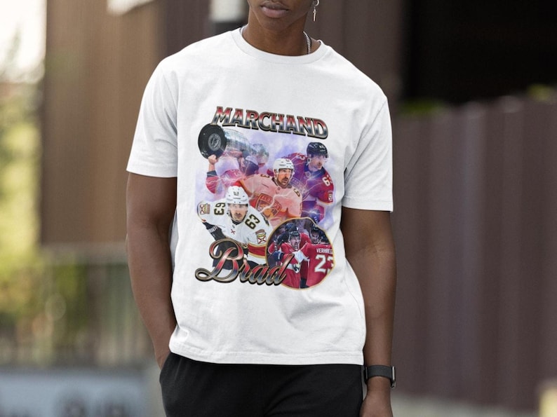 以下が含まれることがあります： 白のTシャツ。ヴィンテージスタイルのグラフィックで、ホッケー選手、上部に「Marchand」、下部に「Brad」の文字。デザインには、番号25の円形画像が含まれています。