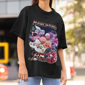 以下が含まれることがあります： 黒いTシャツ。ホッケーをテーマにしたグラフィックデザイン。 「Marchand」と「Brad」の名前、選手とトロフィーの画像が含まれています。 グラフィックはチームカラーを使用し、ダイナミックなスポーツテーマを作成します。