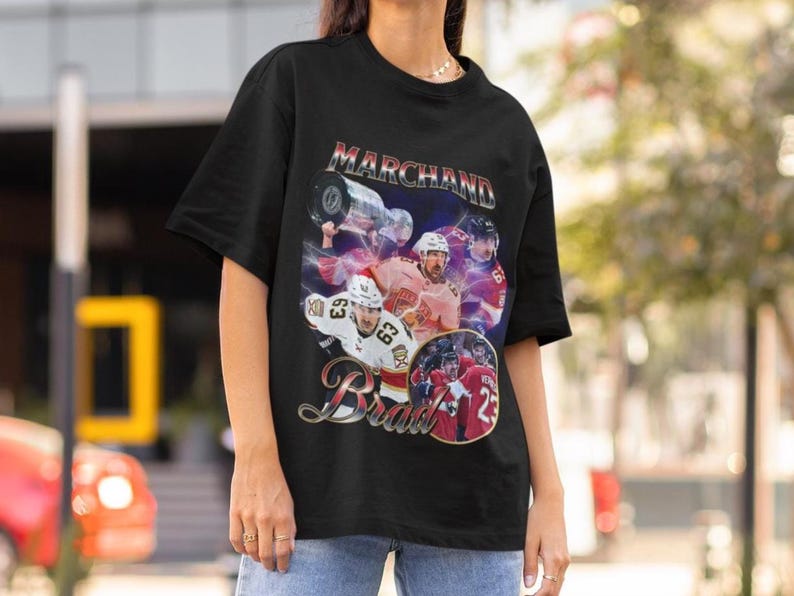 以下が含まれることがあります： 黒いTシャツ。ホッケーをテーマにしたグラフィックデザイン。 「Marchand」と「Brad」の名前、選手とトロフィーの画像が含まれています。 グラフィックはチームカラーを使用し、ダイナミックなスポーツテーマを作成します。