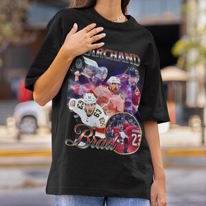以下が含まれることがあります： 黒いTシャツ。ホッケーをテーマにしたデザインで、ホッケー選手のコラージュが特徴です。「Marchand」という名前が上に、「Brad」という名前が下にあります。デザインには、ジャージに63と23の数字が入った選手たちの姿が描かれています。