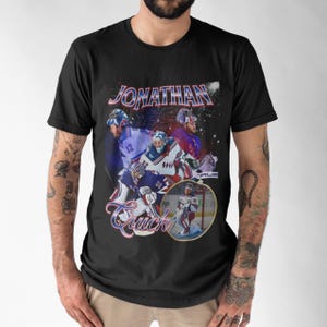 Può includere: T-shirt nero con grafica in stile vintage di un giocatore di hockey. Il design include il nome "Jonathan Quick" in un font stilizzato, insieme a immagini del giocatore in azione. Lo sfondo ha un effetto stellato.