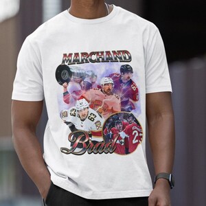 以下が含まれることがあります： 白のTシャツ。ヴィンテージスタイルのグラフィックで、ホッケー選手が描かれています。「Marchand」という名前が上に、「Brad」という名前が下にあります。デザインには、アクション中の選手の画像が含まれており、色あせたレトロな雰囲気です。