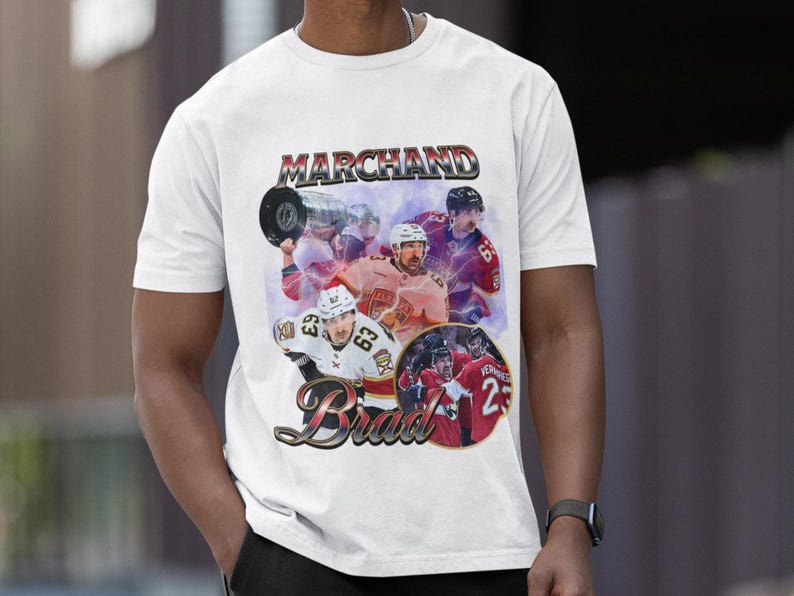 以下が含まれることがあります： 白のTシャツ。ヴィンテージスタイルのグラフィックで、ホッケー選手が描かれています。「Marchand」という名前が上に、「Brad」という名前が下にあります。デザインには、アクション中の選手の画像が含まれており、色あせたレトロな雰囲気です。