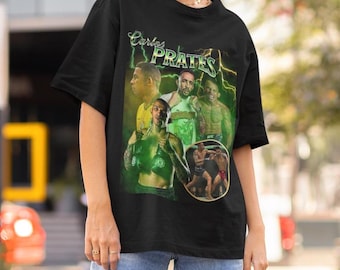 May include: A black short-sleeve t-shirt featuring a graphic print of fighter Carlos Prates. The design includes multiple images of the athlete, green lightning accents, and the text Carlos Prates in a stylized font across the chest area.