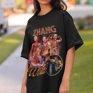 May include: Black t-shirt with a graphic of mixed martial artist Zhang Weili. The design includes multiple images of Zhang, with the name "ZHANG" in gold lettering at the top and "Weili" in gold script at the bottom.