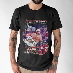 以下が含まれることがあります： 黒いTシャツ。ヴィンテージスタイルのグラフィックで、ホッケー選手「Marchand」と「Brad」の名前が装飾的なフォントで描かれています。デザインには、紫と赤の背景の中で、選手が活躍している様子が描かれています。
