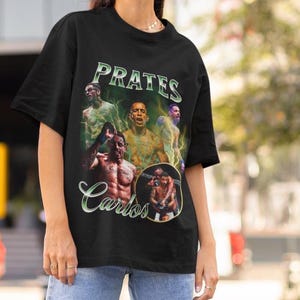 May include: Black t-shirt featuring a graphic design with the name "PRATES" in green and white, and the name "Carlos" in white script. The design includes images of several men in action poses, with a vintage aesthetic.