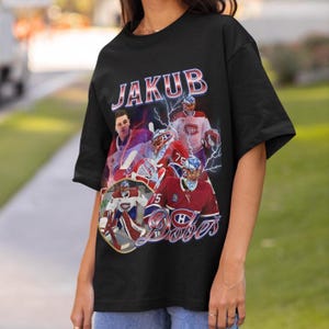 Op de afbeelding: Een zwart T-shirt met korte mouwen en een grafische print van ijshockeykeeper Jakub Dobeš. Het ontwerp bevat meerdere afbeeldingen van de speler in een rood-wit shirt, blauwe bliksemschichten en de tekst JAKUB DOBEŠ in vetgedrukte letters.