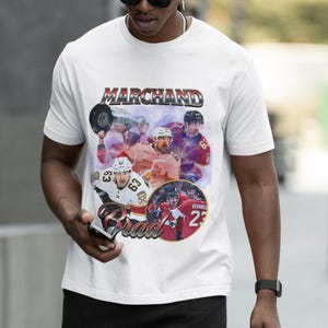 以下が含まれることがあります： 「MARCHAND」の文字が銀と赤で描かれたグラフィックデザインの白いTシャツ。ホッケー選手の画像が含まれています。カジュアルウェアアイテムです。