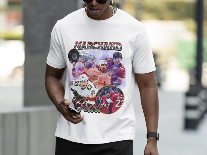 以下が含まれることがあります： 「MARCHAND」の文字が銀と赤で描かれたグラフィックデザインの白いTシャツ。ホッケー選手の画像が含まれています。カジュアルウェアアイテムです。
