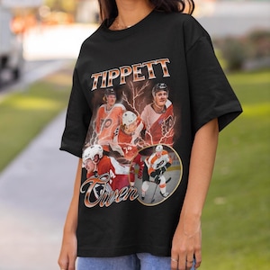 Könnte beinhalten: Schwarzes T-Shirt mit einem Vintage-Design, das einen Eishockeyspieler, den Namen "Tippett" in Orange und Weiß sowie Bilder des Spielers mit Blitzeffekten zeigt. Ein Sport-Themen-Kleidungsstück.