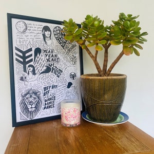 May include: A black and white framed print with a hand-drawn illustration of a lion, a woman, and a variety of text. The text includes the words "Yeah Yeah Yeahs", "Off Off", "Dance Dance Dance", and "Till You're Dead". The print is sitting on a wooden table next to a potted jade plant and a pink candle.