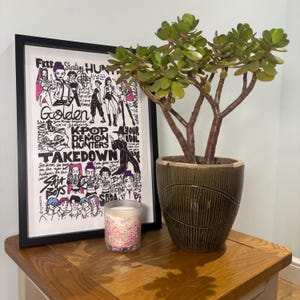 May include: A black framed art print with the words "Golden", "KPOP DEMON HUNTERS", and "TAKEDOWN". A jade plant in a brown pot and a candle with pink flecks are on a wooden table.