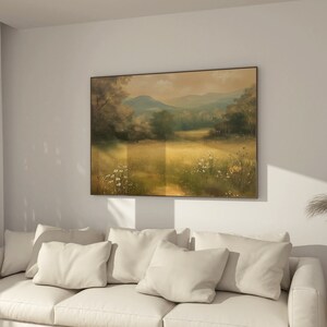 May include: A white couch with three pillows in a living room with a framed landscape painting of a field with trees and mountains in the background.