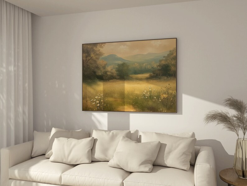 May include: A white couch with three pillows in a living room with a framed landscape painting of a field with trees and mountains in the background.