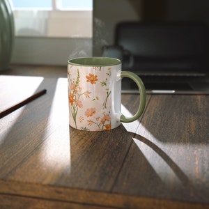 May include: A white ceramic mug with a floral pattern in shades of orange, pink, and green. The mug has a green rim and handle. Steam is rising from the mug, suggesting it is filled with a hot beverage.