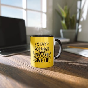 May include: Yellow coffee mug with a black rim and handle. The mug has black text that reads "Stay Focused and Never Give Up".
